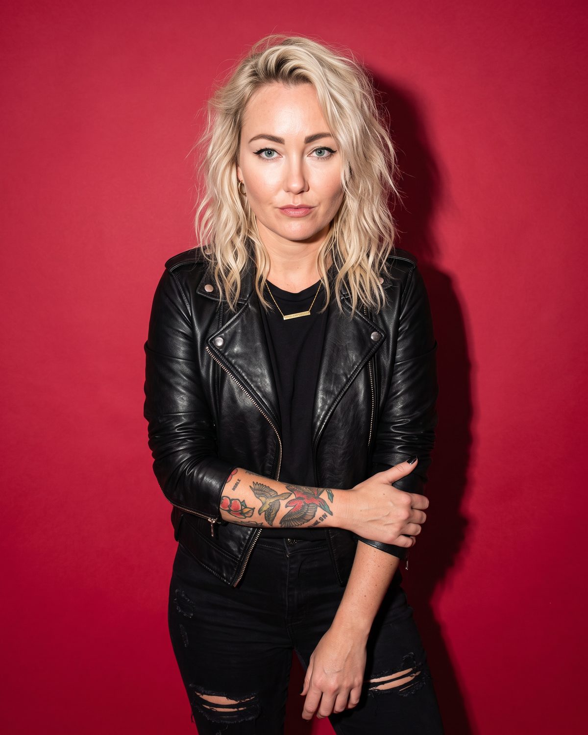 Jessie Riot against a cherry-red studio backdrop, arms folded, in a black leather jacket and ripped black jeans.