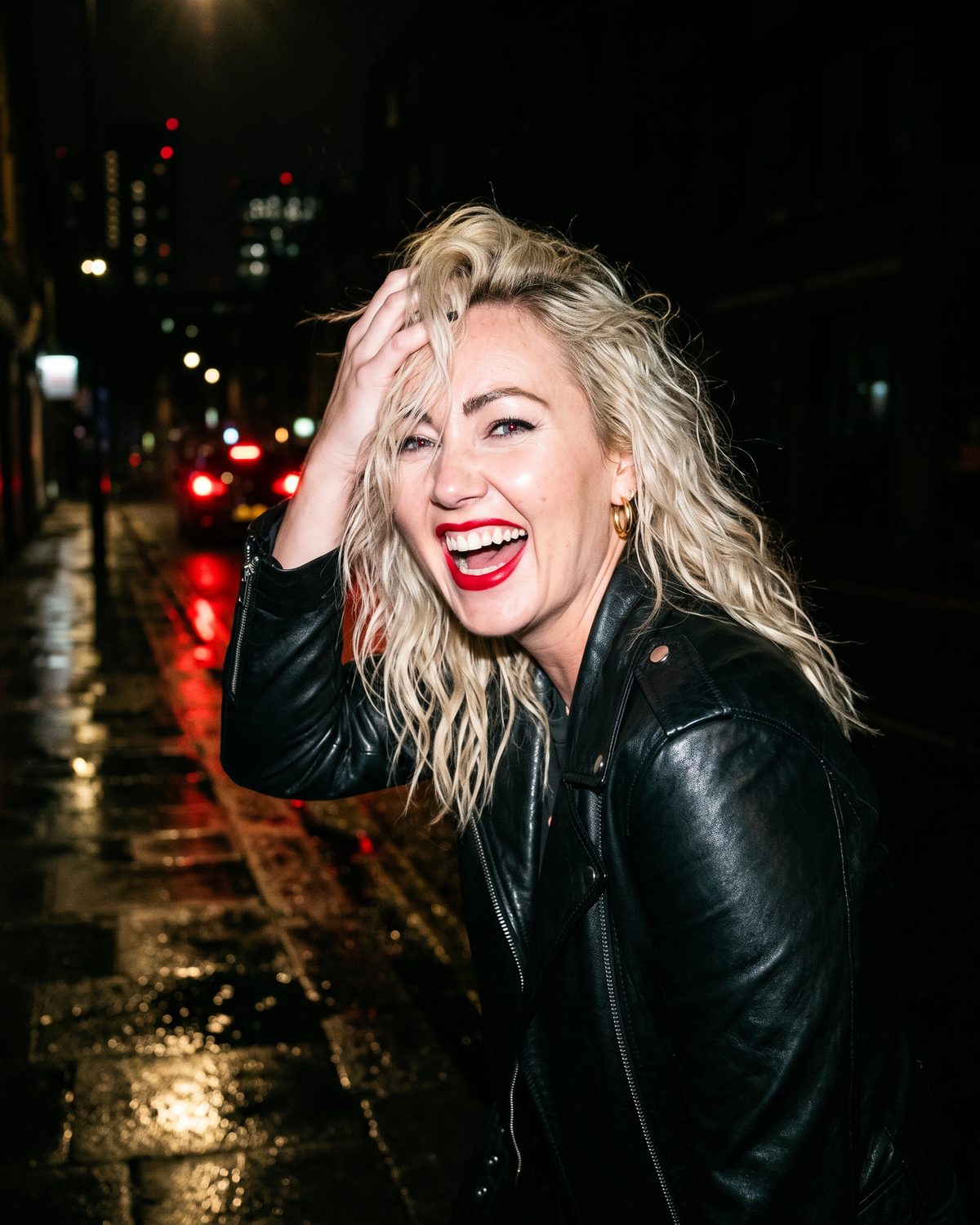Jessie Riot laughing on a wet street at night, lit by a camera flash, pushing her hair back, red tail lights behind her.