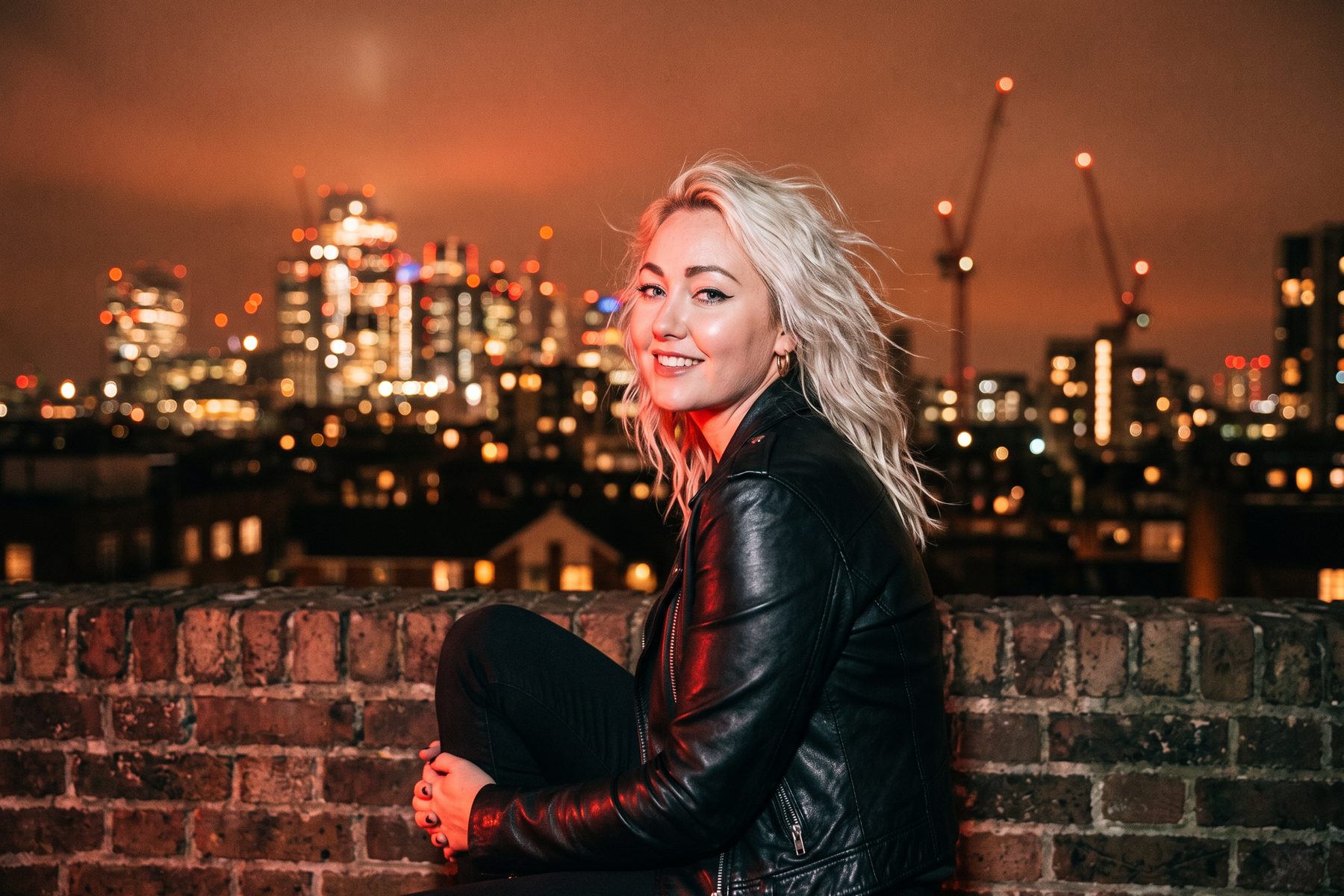 Jessie Riot sitting on a brick rooftop wall at night, grinning over her shoulder, with the lights of the London skyline blurred behind her.
